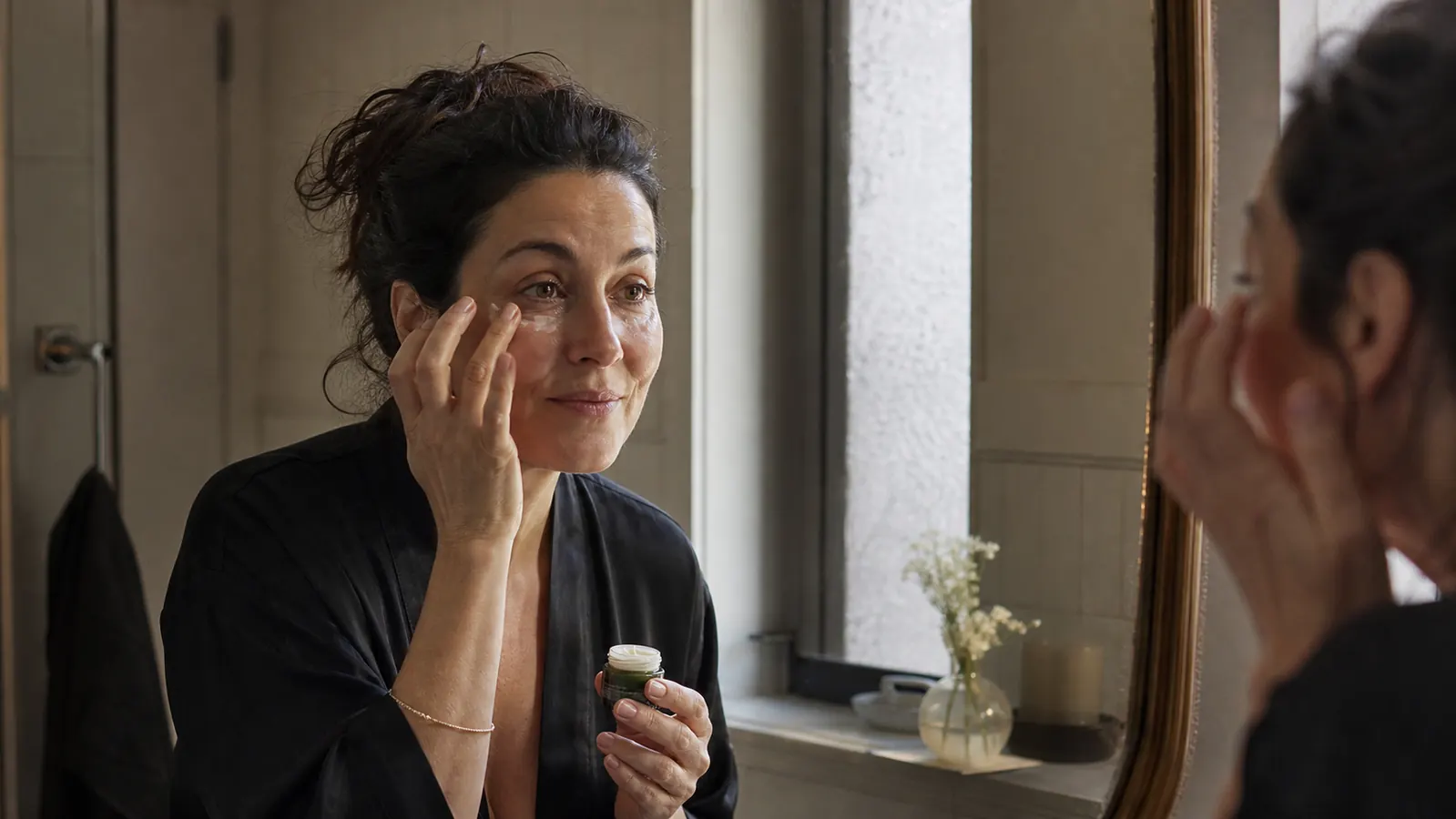 Woman in her 40s at a bathroom mirror in soft morning light, dabbing eye cream under her eyes with her ring finger