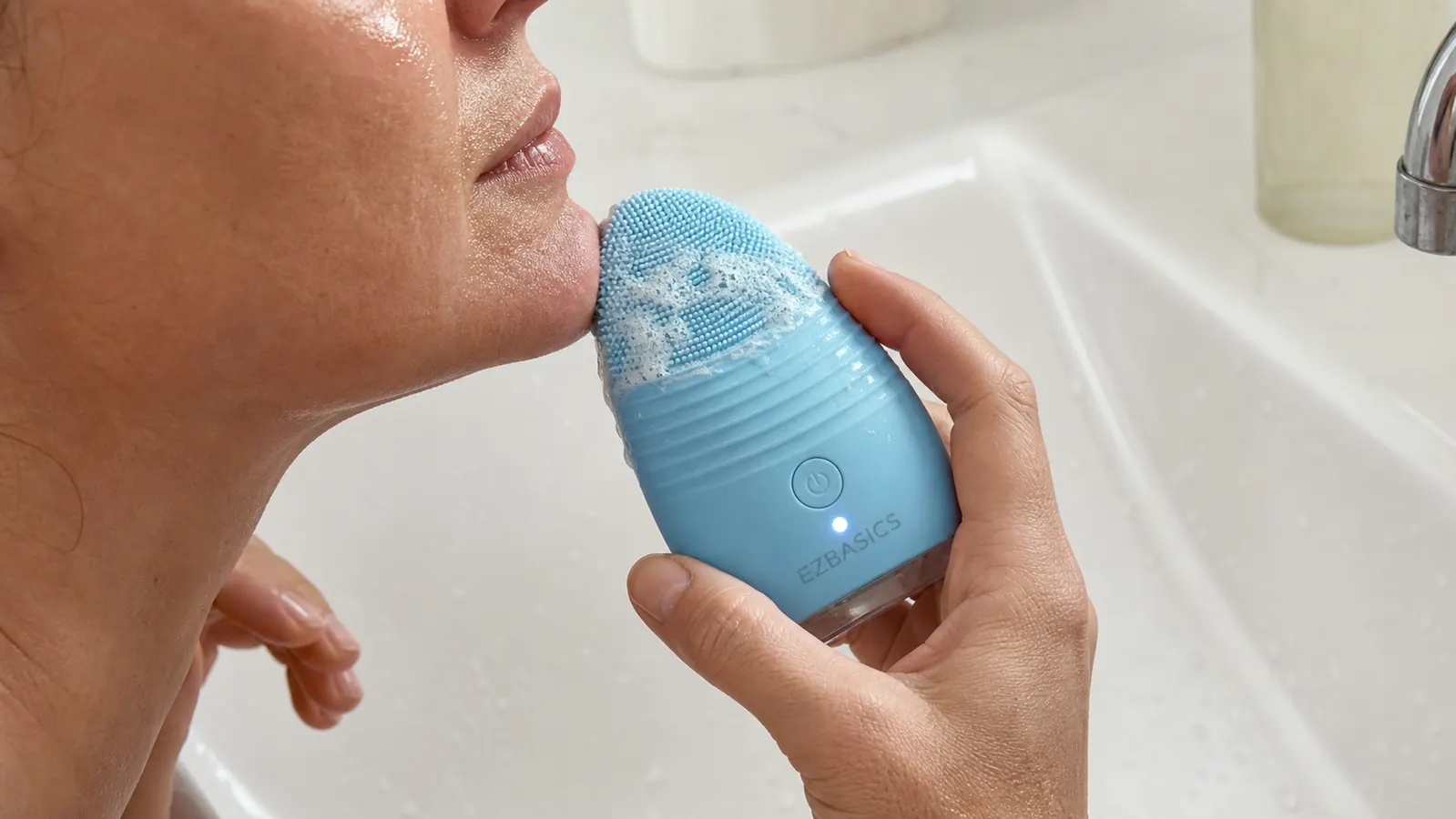 Close-up of a hand holding a silicone facial cleansing brush with light foam, rinsing at a bathroom sink