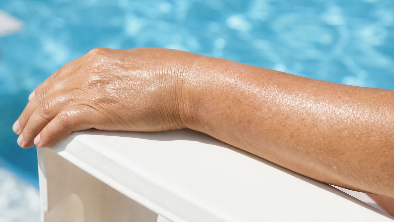 Close-up of a woman's bare, sunscreen-covered forearm resting on a pool railing on a bright day