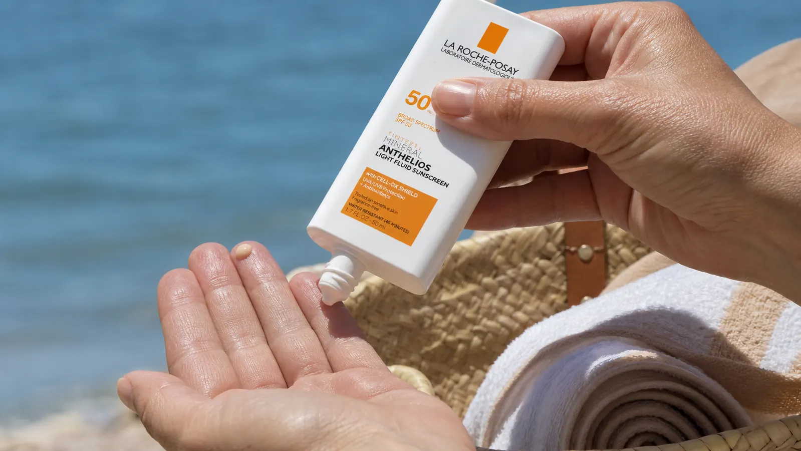 Close-up of a hand squeezing a small amount of tinted sunscreen fluid onto fingertips beside a beach bag and towel