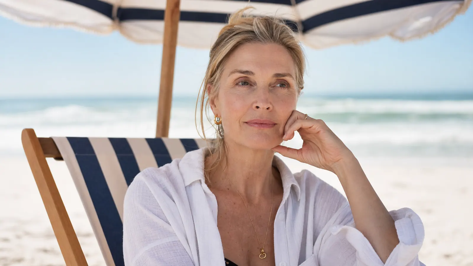 Woman in her 50s sitting under a beach umbrella, gently touching her cheek and looking out at the water, calm expression with no visible redness or shine