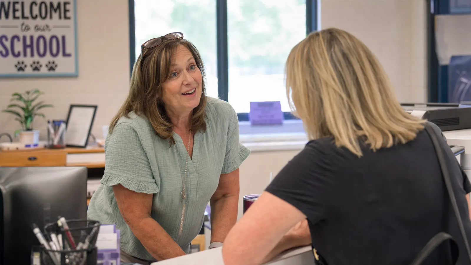 Two women chatting and smiling at a school front desk in bright morning light