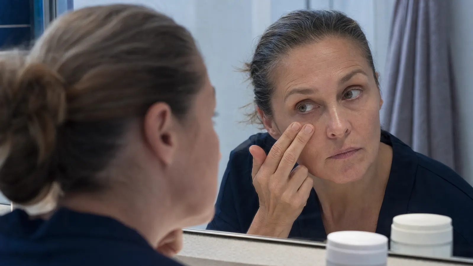 A woman in her late forties leaning close to a bathroom mirror in cool morning light, examining the skin under her eyes with two fingers