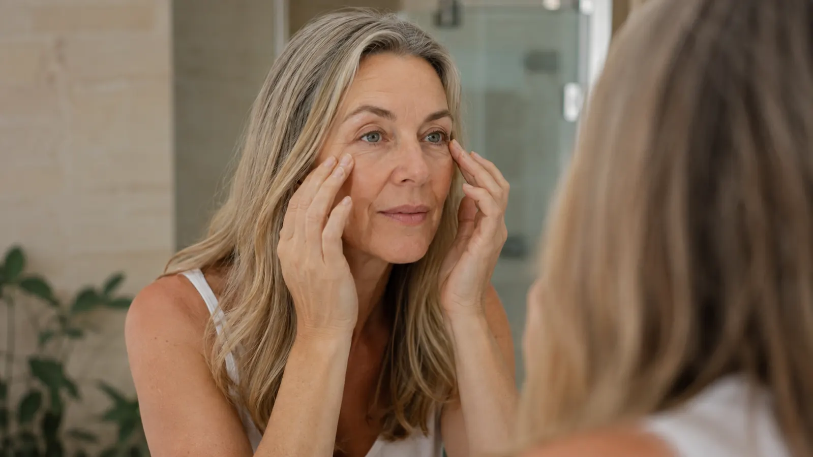 Woman in her 50s in a bright bathroom mirror, morning light, gently pressing under her eyes with clean fingertips before applying any product