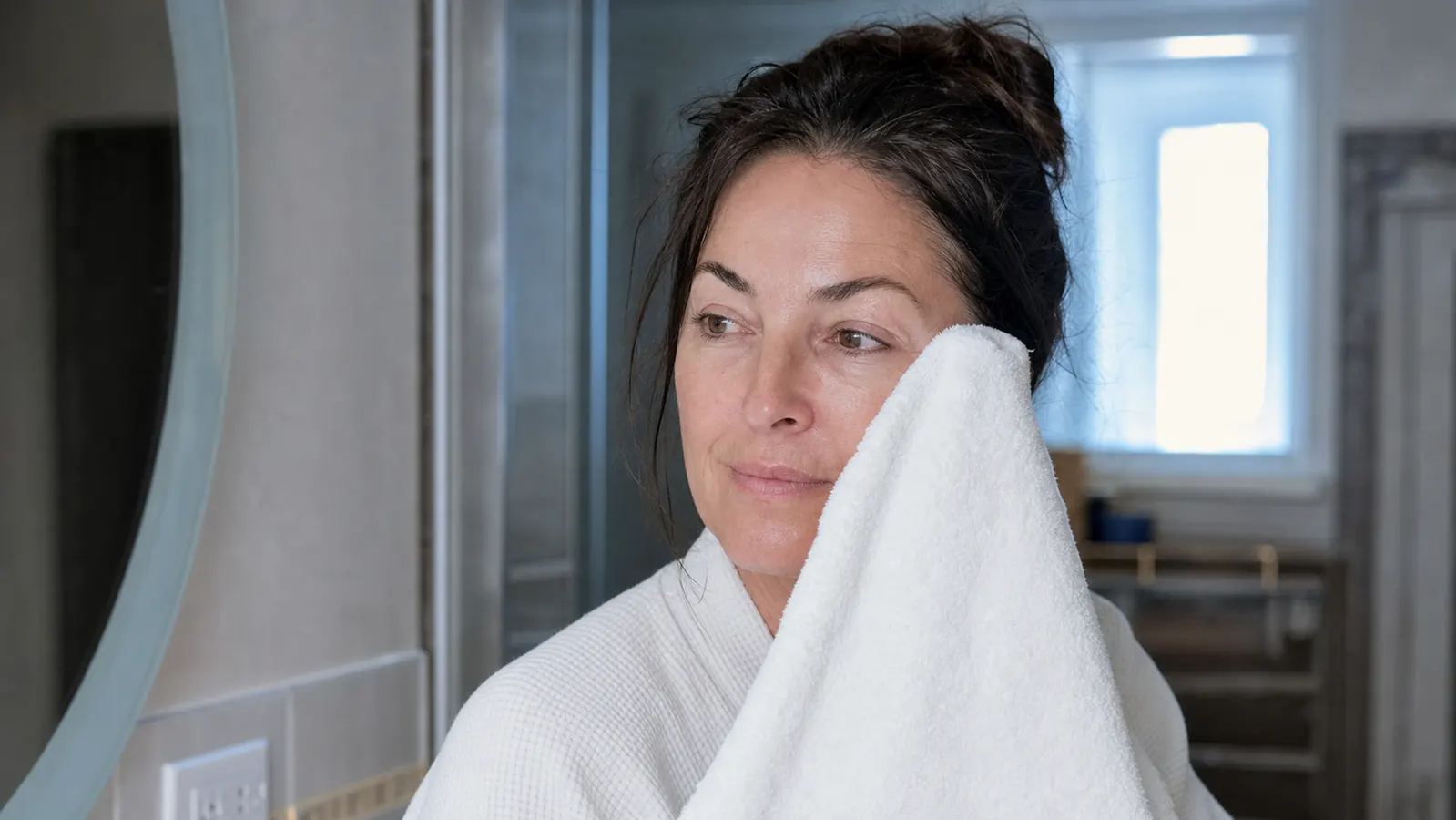 Woman in her late 40s leaning over a bathroom sink, patting her face dry with a white towel, morning light coming through a small window